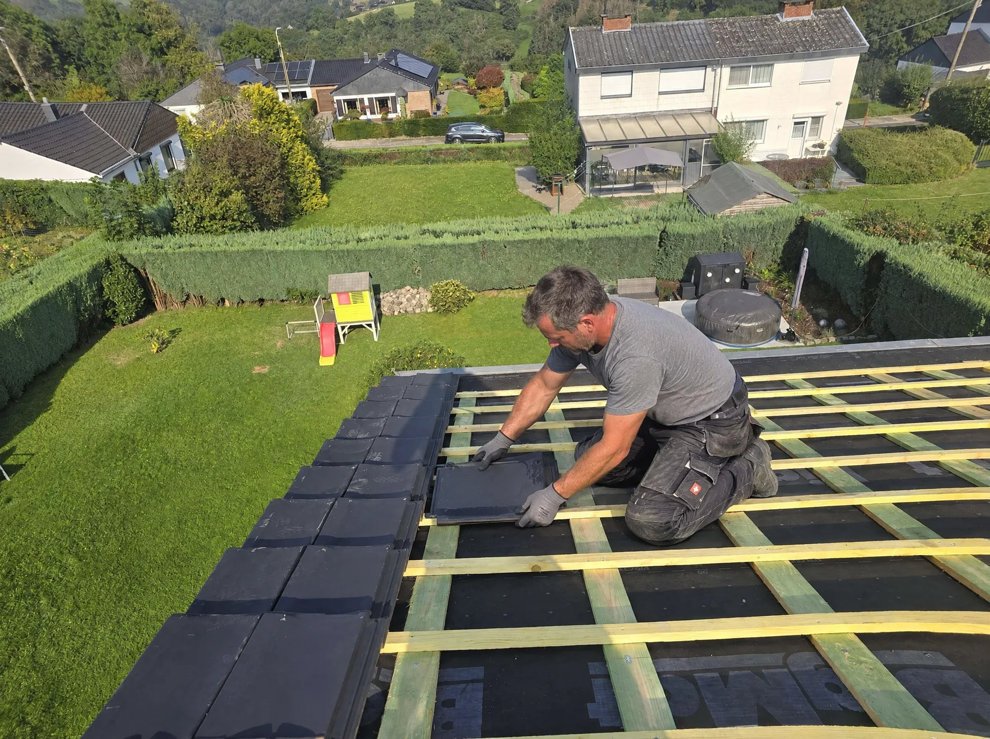 Un couvreur pose une tuile sur un lattage neuf, vue sur le jardin et les maisons voisines